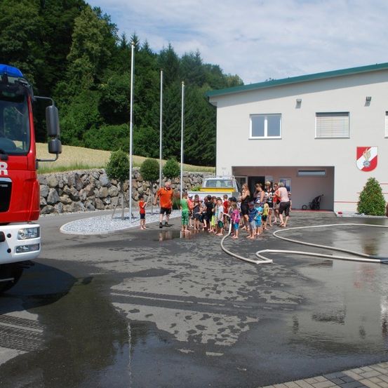 Eine Gruppe von Kindern steht vor einer Feuerwache mit einem roten Feuerwehrauto und einer Feuerwehrhose. Das Gebäude hat ein Logo an der Wand.