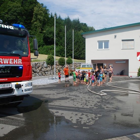 Eine Gruppe von Kindern steht in einer Reihe und beobachtet einen Feuerwehrwagen, mit einer Feuerwache im Hintergrund.