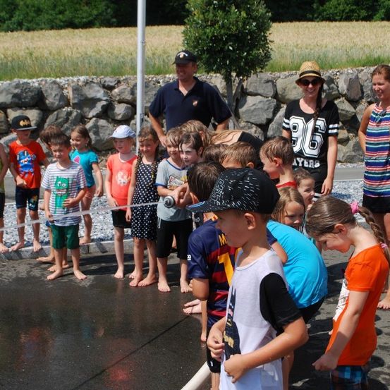 Kinder stehen auf nassem Bürgersteig und halten einen Wasserschlauch, umgeben von Erwachsenen. Ein großer Steinwall steht hinter ihnen, und ein Feld ist im Hintergrund.