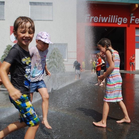 Mehrere Kinder spielen unter einem Sprinkler vor einem roten Gebäude mit der Aufschrift 'Freiwillige Feuer'. Sie lächeln und scheinen das Wasser zu genießen.