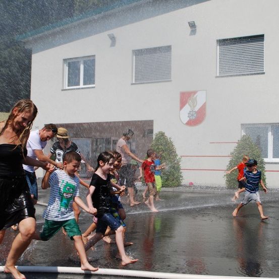 Mehrere Kinder und Erwachsene spielen draußen im Regen, in der Nähe eines Gebäudes mit einem Logo an der Wand.