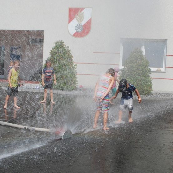 Kinder spielen vor einem Gebäude mit Logo im Wasser, das aus einer Fontäne spritzt.