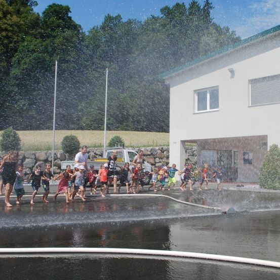 Kinder spielen in einem Wasserbrunnen unter einem strahlenden Himmel. Dahinter steht ein Gebäude mit einem geparkten Pickup-Truck.