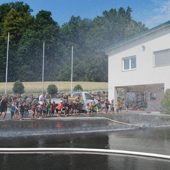 Eine Gruppe von Kindern und Erwachsenen spielt in einem Wassersprüher vor einem Gebäude, mit einem weißen Pickup-Truck in der Nähe und Bäumen im Hintergrund.