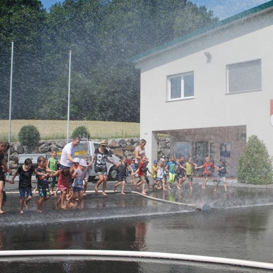 Kinder spielen in einer Wasserfontäne vor einem Gebäude mit einem Feuerwehrmann-Emblem an der Wand.