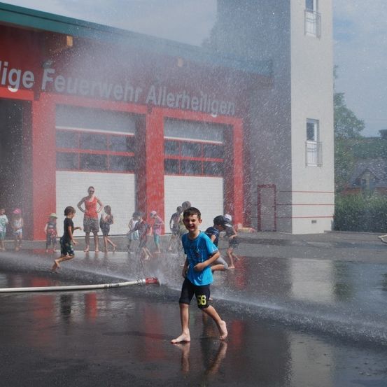 Ein Junge spielt unter einem Wasserschlauch vor einer Feuerwache, umgeben von anderen Kindern.