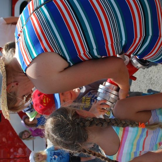 Eine Frau und ein Mädchen stehen neben einem Feuerwehrauto. Die Frau hält eine Dose und das Mädchen schaut sie an. Hinter ihnen stehen Kinder, von denen einige Mützen tragen.