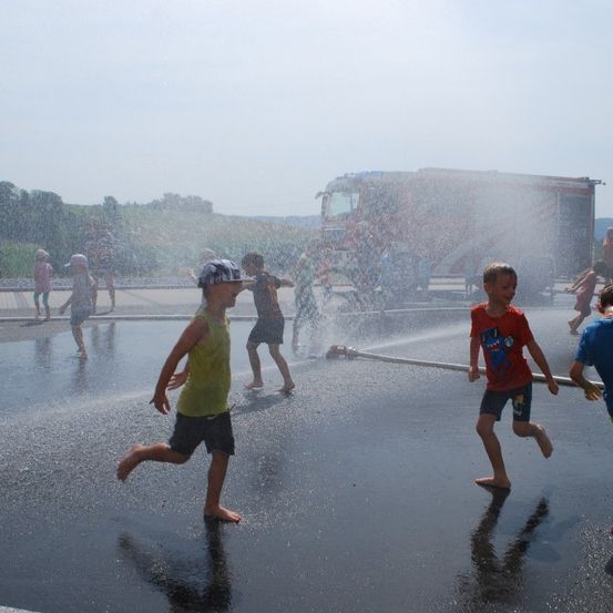 Mehrere Kinder laufen an einem sonnigen Tag durch einen Wasserstrahl eines Feuerwehrwagens. Einige Erwachsene stehen in der Nähe.