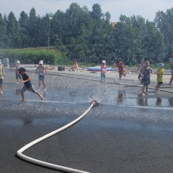 Kinder spielen mit Wasser, das aus einem Schlauch gesprüht wird, an einem sonnigen Tag. Bäume und ein Gebäude sind im Hintergrund.