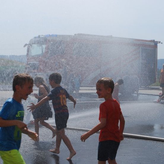 Eine Gruppe von Kindern spielt mit dem Wasserschlauch eines Feuerwehrwagens, mit einem klaren Himmel im Hintergrund.