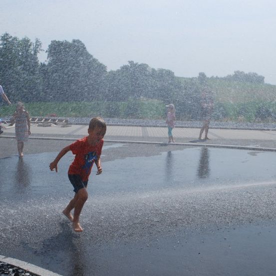 Mehrere Kinder und Erwachsene genießen einen nassen Tag in einem Wasserpark. Ein Junge läuft durch einen Wasserbrunnen.