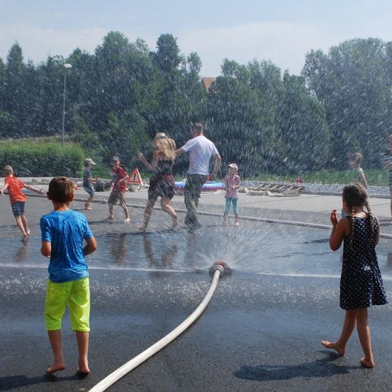 Eine Gruppe von Kindern und Erwachsenen spielt in einem Wasserbrunnen in einem Außenbereich mit Bäumen und einem Gebäude im Hintergrund.