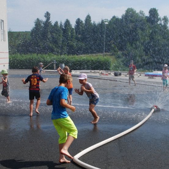 Mehrere Kinder spielen an einem sonnigen Tag in einem Wasserbrunnen, mit Bäumen und einem Gebäude im Hintergrund.