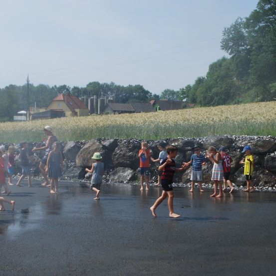 Kinder spielen auf einer nassen Straße, in der Nähe von Felsen und einem Weizenfeld. Einige Erwachsene sind in der Nähe. Bäume und Häuser sind im Hintergrund.