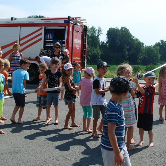 Mehrere Kinder und Erwachsene stehen vor einem Feuerwehrauto und halten eine lange Feuerwehrschläuche. Einige der Kinder tragen Mützen. Die Erwachsenen beobachten die Kinder.