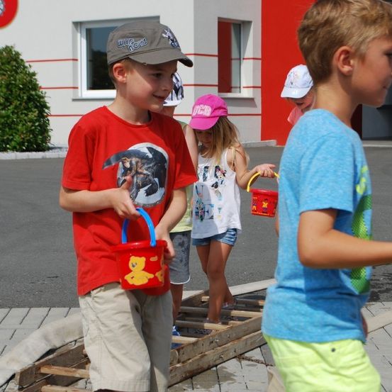 Eine Gruppe von Kindern läuft draußen auf einer hölzernen Rampe. Sie halten rote Eimer. Ein Gebäude steht hinter ihnen.