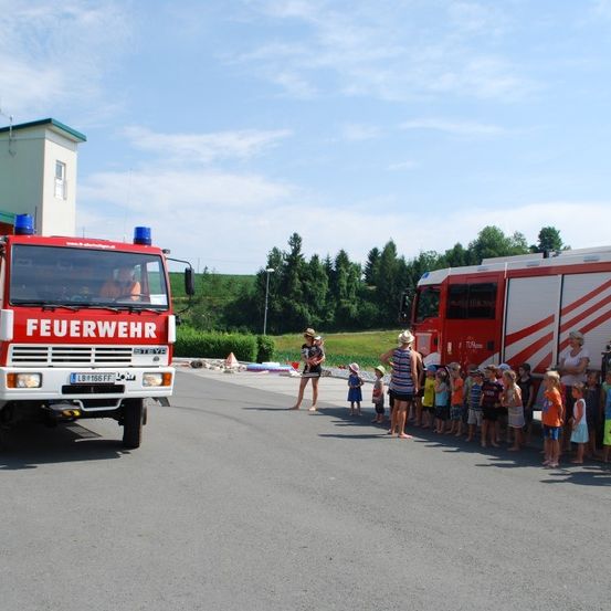Eine Reihe von Kindern und Erwachsenen steht neben zwei roten Feuerwehrautos mit anwesenden Feuerwehrleuten an einem sonnigen Außenbereich.