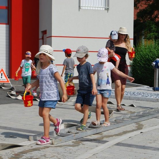 Eine Gruppe von Kindern mit Hüten und Eimern in der Hand geht auf einem nassen Pfad vor einem Gebäude mit rot-weißer Wand.