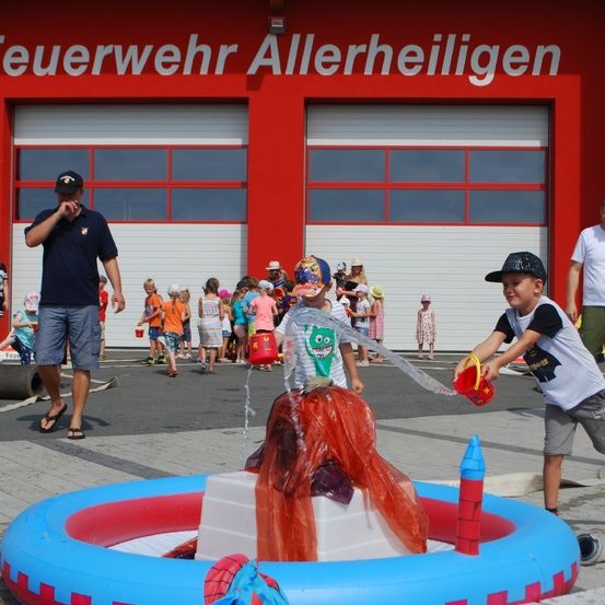Ein junger Junge spritzt Wasser aus einem Eimer auf eine Wasserfontäne in der Feuerwache Allerheinligen. Hinter ihm steht ein Mann und beobachtet. Viele Kinder stehen in einer Reihe und warten auf ihren Zug.