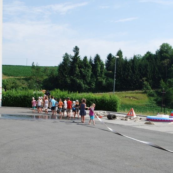 Mehrere Kinder stehen in einem Parkplatz und halten ein langes Band. In der Nähe ist ein Warnschild auf dem Boden platziert. Dahinter ist ein Feld und Bäume zu sehen.