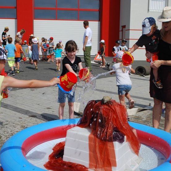 Kinder spielen mit Wasser in einem großen Pool. Eine Erwachsene hilft ihnen, Wasser aus Eimern zu gießen. In der Nähe spielen auch andere Kinder mit Wasser und andere stehen herum.