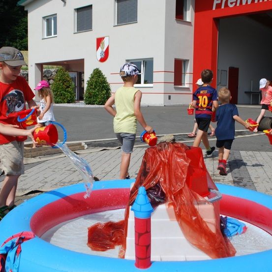 Kinder spielen um einen aufblasbaren Pool mit Wasser und halten Eimer. Dahinter ist ein Gebäude mit einem Feuerwehrlogo zu sehen.