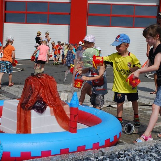 Kinder spielen in einem bunten aufblasbaren Pool und halten Eimer. Ein Kind gießt Wasser auf einen roten Stoff. Ein Mann in einem blauen Hemd steht hinter ihnen.