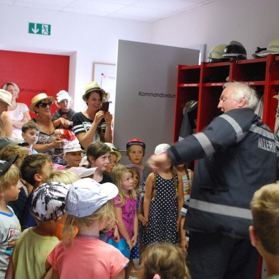 Ein Feuerwehrmann in Uniform spricht zu einer Gruppe von Kindern in einem Raum mit roten Schließfächern und Helmen. Einige Erwachsene sind anwesend, darunter eine Frau, die ein Foto macht.