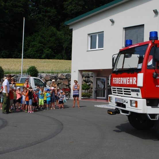 Eine Gruppe von Kindern und Erwachsenen hat sich vor einem Feuerwehrauto auf einem offenen Gelände versammelt. Das Feuerwehrauto trägt den Schriftzug 'Feuerwehr' an seiner Seite. Dahinter befindet sich ein Gebäude mit Glasfenstern und Türen.