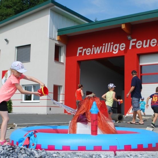 Kinder spielen mit Wasser vor einem Feuerwehrhaus. Ein Mädchen gießt Wasser aus einem Eimer.