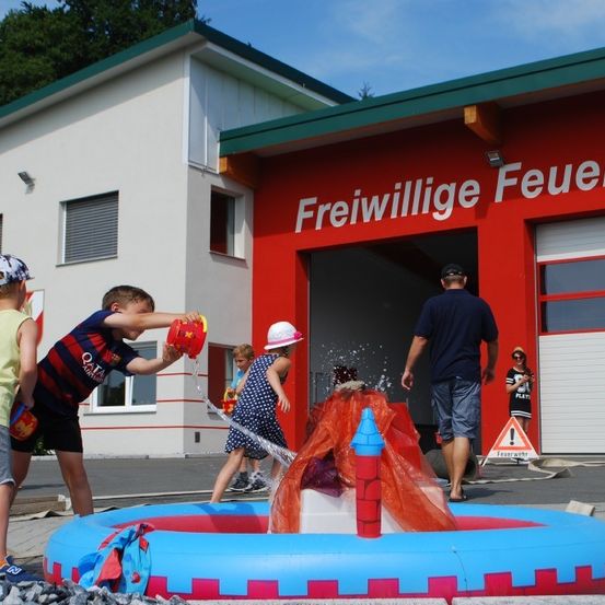 Kinder spielen mit Wassereimern vor einer Feuerwehrstation, wobei ein Kind einen Helm und ein anderes einen Hut trägt. Ein Mann geht an ihnen vorbei.