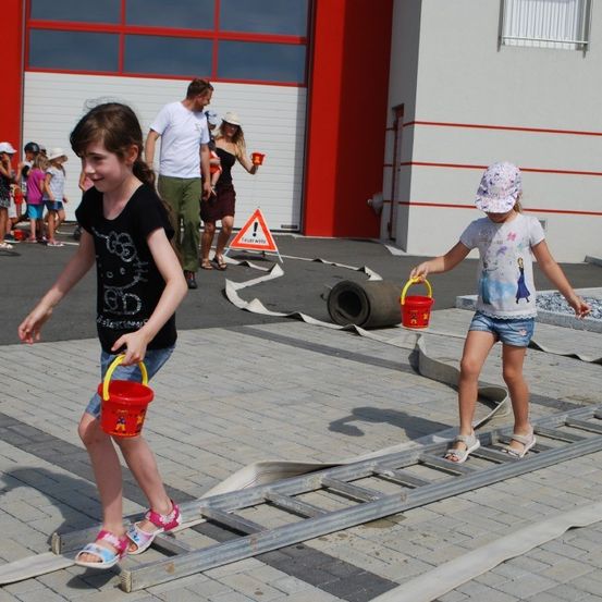 Zwei Mädchen gehen mit Eimern in der Hand über eine Leiter draußen, Erwachsene und Kinder stehen in der Nähe, ein Gebäude mit roten Säulen ist im Hintergrund.