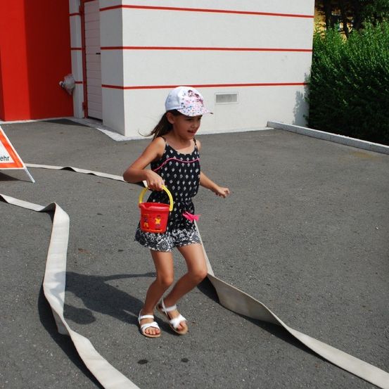 Ein kleines Mädchen mit einem Eimer in der Hand geht auf einem mit weißen Bändern markierten Pfad auf einer Betonfläche. Ein dreieckiges Warnschild ist links aufgestellt.