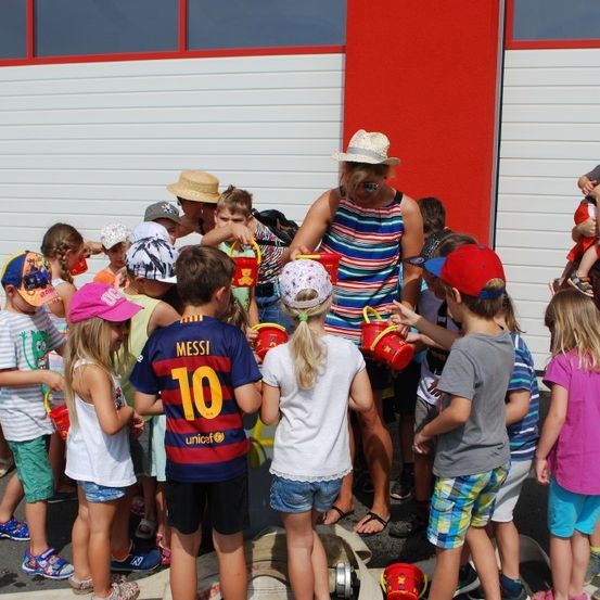 Kinder versammeln sich um Erwachsene, einige halten rote Eimer. Eine Erwachsene gibt den Kindern Eimer aus, und andere beobachten. Die Umgebung scheint im Freien zu sein, mit einem Gebäude im Hintergrund.