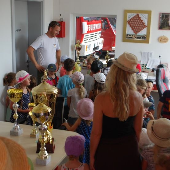 Eine Gruppe von Kindern und Erwachsenen versammelt sich um einen Tisch mit Trophäen. Ein Mann steht hinter ihnen, und ein Feuerwehrauto ist durch eine Glastür sichtbar.