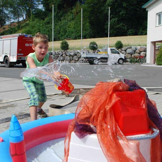 Ein junger Junge spielt draußen mit einer Wasserpistole. Er steht vor einem aufblasbaren Pool mit einem roten Eimer. Dahinter sind ein Feuerwehrauto und ein weißer Lastwagen geparkt. Bäume und ein Haus sind im Hintergrund.