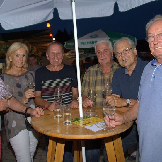 Fünf Personen sitzen bei Nacht unter einem Schirm um einen Tisch und halten Weingläser. Sie scheinen sich auf einer gesellschaftlichen Veranstaltung zu befinden. Eine Frau auf der linken Seite hält ein Weinglas und eine Speisekarte.