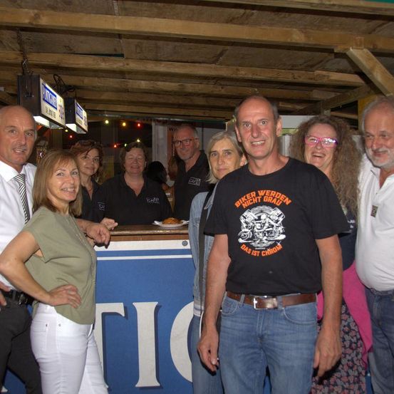 Eine Gruppe von Erwachsenen steht in einer Bar und lächelt für die Kamera. Eine Frau in Grün lehnt sich an eine Theke, während ein Mann in einem schwarzen T-Shirt einen Gürtel trägt.