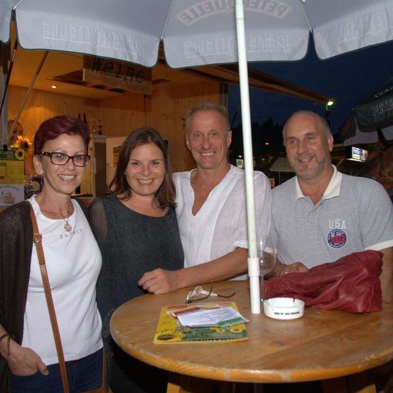 Vier Personen, zwei Männer und zwei Frauen, lächeln und posieren für ein Foto an einem Tisch unter einem großen Schirm. Der Tisch hat einen weißen Stab, ein Glas und eine rote Tasche. Dahinter steht eine Holzkonstruktion mit einem Schild und einem Regal mit Flaschen.