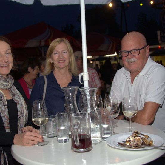 Drei Personen sitzen um einen Tisch mit Weinglasern und einer Dessertplatte. Der Mann hat einen Schnurrbart und eine Brille. Die Frau links trägt einen Schal. Hinter ihnen sind Zelte und Menschen.