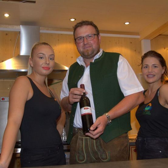 Drei Personen in einer Küche, ein Mann hält eine Flasche Wein. Zwei Frauen stehen auf beiden Seiten von ihm.