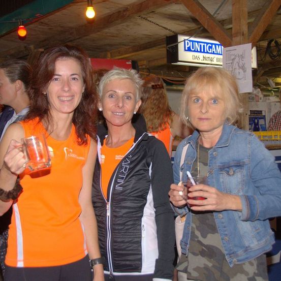 Drei Frauen stehen zusammen drinnen und lächeln in die Kamera. Die Frau auf der linken Seite trägt ein orangefarbenes Shirt und hält ein Glas. Die Frau in der Mitte trägt eine schwarze Jacke. Die Frau rechts trägt eine Jeansjacke und hält einen Becher. Hinter ihnen steht ein Schild mit der Aufschrift 'PUNTIGAM'.