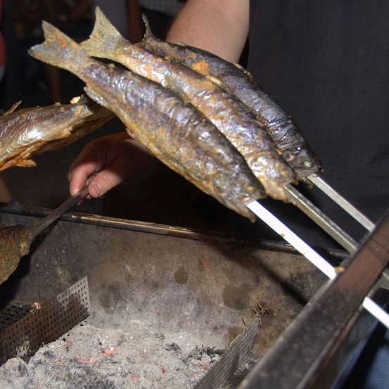 Eine Person grillt Fisch auf einem Grill im Freien. Sie hält ein Messer und die Fische sind auf Spießen. Die Fische sind gekocht und der Grill ist rauchig.