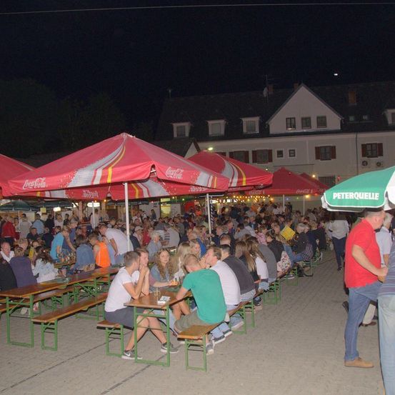 Ein überfüllter Außenbereich mit mehreren Tischen und Bänken unter Coca-Cola-Schirmen. Menschen sitzen und stehen, einige gehen. Ein Gebäude mit Giebeldach ist im Hintergrund.