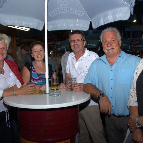 Vier Personen posieren für ein Foto draußen und halten Gläser mit Bier in der Hand. Hinter ihnen steht ein Schirm mit dem Wort "Hefeteig".