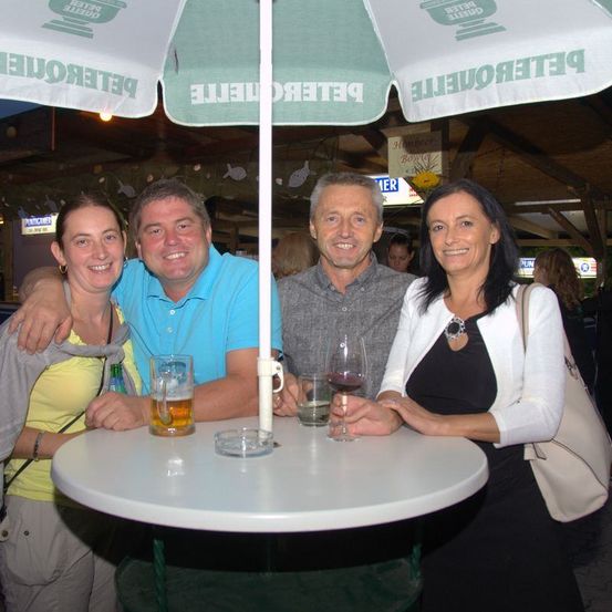 Vier Personen, zwei Männer und zwei Frauen, lächeln und posieren für ein Foto. Sie sitzen an einem Tisch unter einem grünen Schirm. Sie halten Gläser mit Bier und Wein.