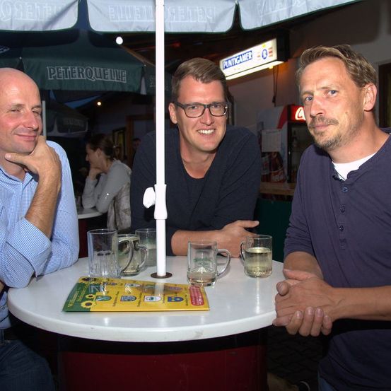 Drei Männer sitzen an einem Tisch mit Getränken, zwei mit Brillen. Dahinter ein Regenschirm, ein Coca-Cola-Kühler und ein Schild mit der Aufschrift Peterquelle.