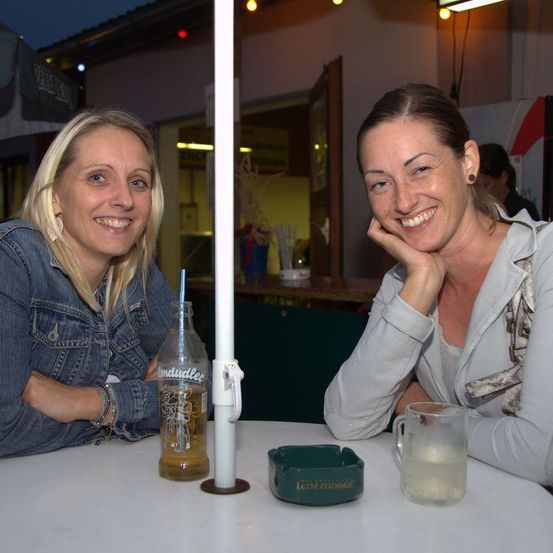 Zwei Frauen lächeln und posieren für ein Foto in einem Restaurant im Freien, mit einer Flasche Bier, einem Glas Flüssigkeit und einem Aschenbecher auf dem Tisch.