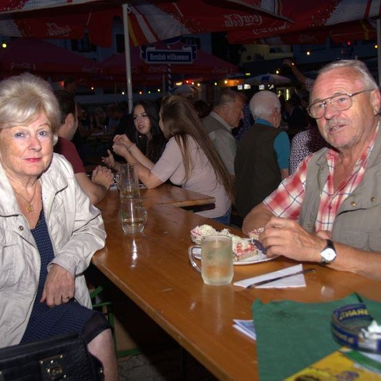 Eine Gruppe älterer Menschen isst in einem Restaurant im Freien. Sie sitzen um einen Holztisch herum mit Tellern mit Essen und Gläsern mit Getränken.