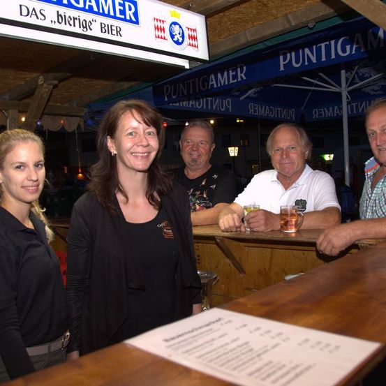 Vier Menschen lächeln an einem Tresen. Eine Speisekarte liegt auf dem Tresen und über ihnen steht ein Schild mit der Aufschrift Das Bierige Bier.